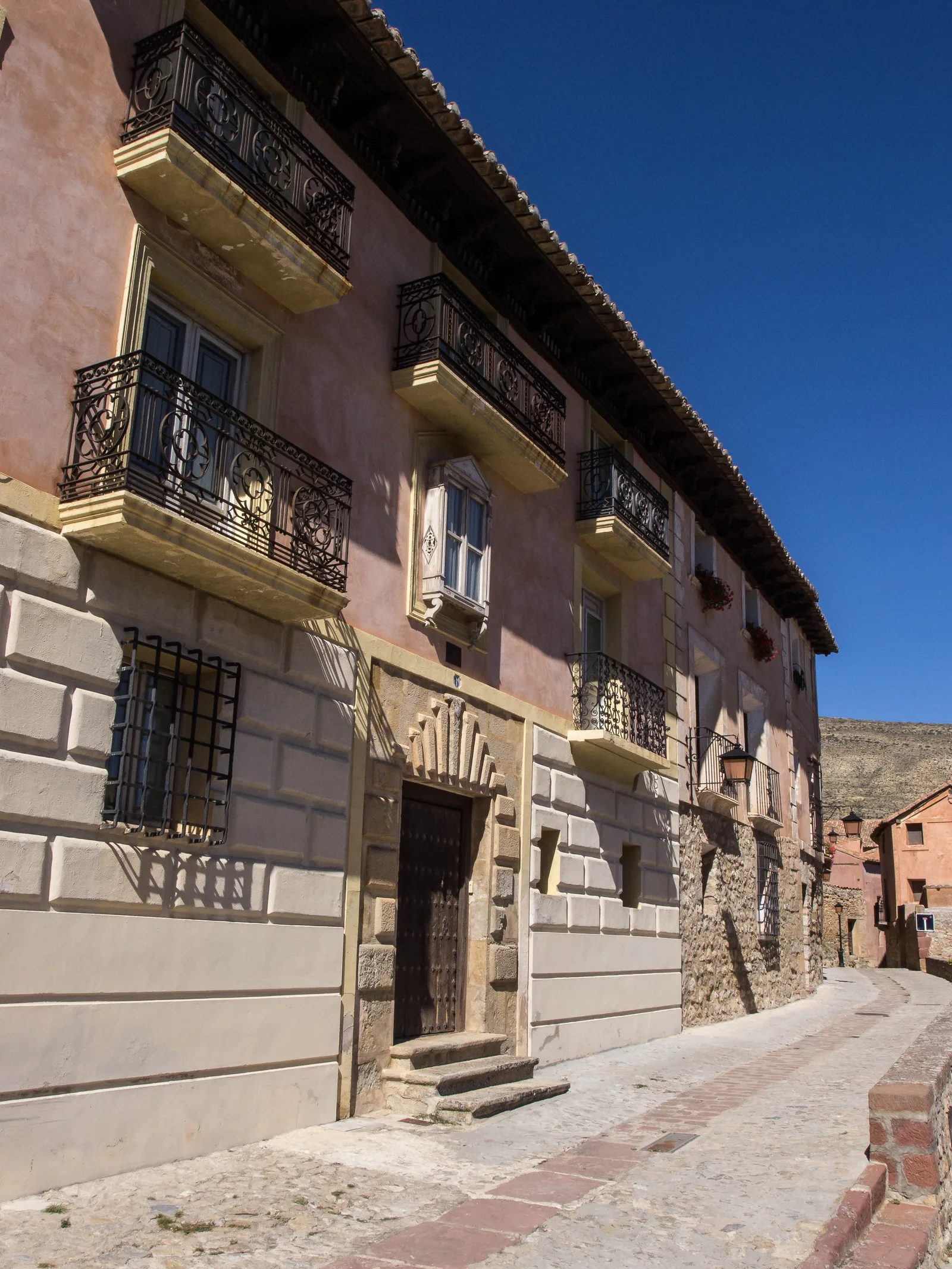 A narrow paved lane beside a pink building with wrought-iron balconies and a stone doorway.