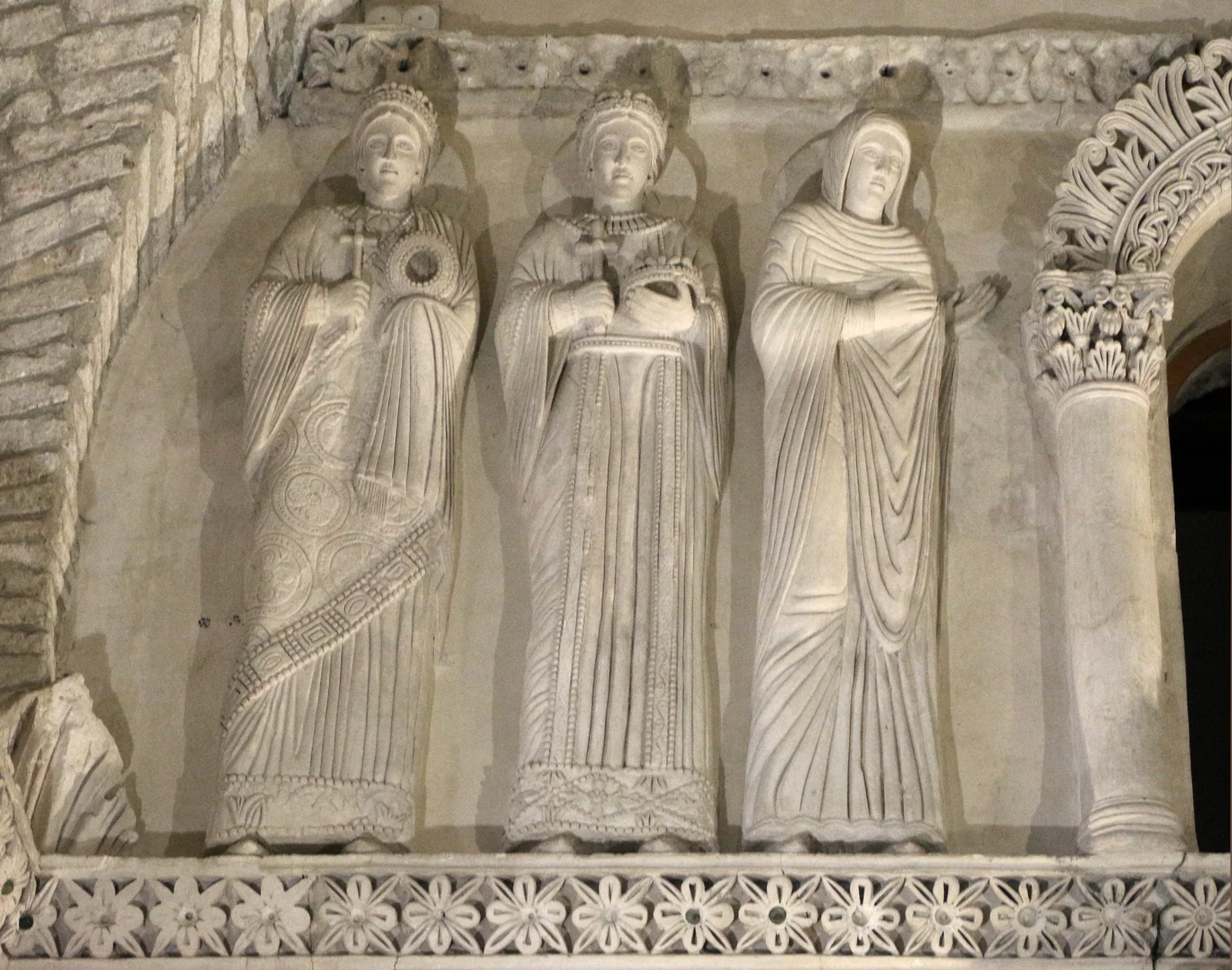 Three haloed female stucco figures in long robes beside carved ornament inside the Tempietto Longobardo.