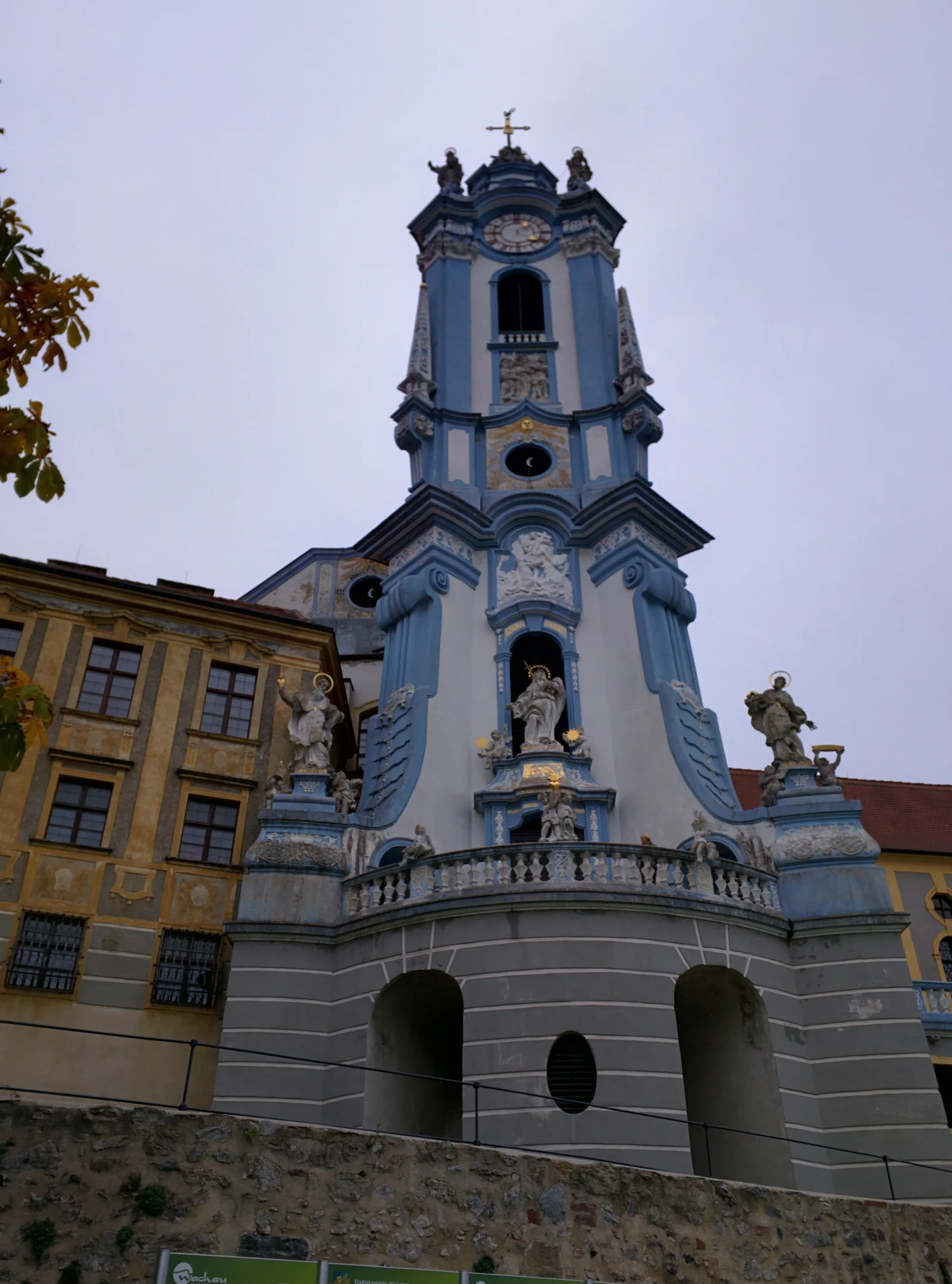 The blue-and-white tower of Dürnstein Abbey seen from below, with a clock, sculpted figures and an adjoining yellow building.